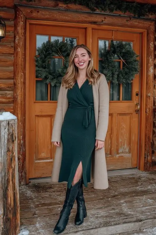 A woman over 30 wears a dark green midi sweater dress with long sleeves and a slit, paired with tall black cowboy boots and a camel wrap coat