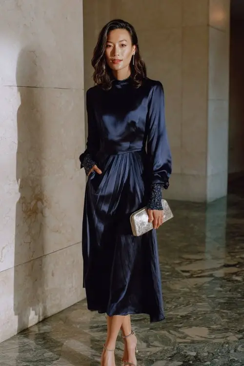 A woman over 30 wears a long-sleeve navy satin dress with embroidered cuffs and a pleated skirt