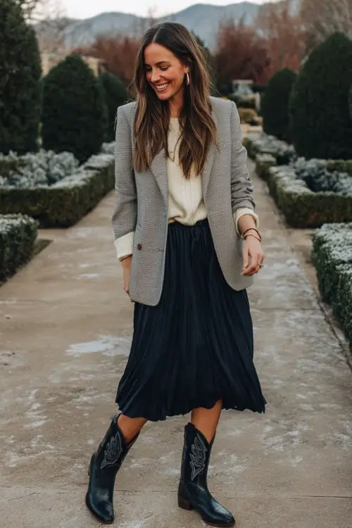 A woman over 30 wears a navy pleated midi skirt with a tucked-in cream knit, layered with a gray wool blazer and tall black cowboy boots