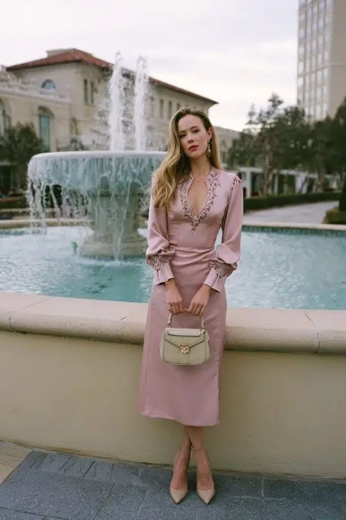A woman over 30 wears a rose-colored midi dress with full-length sleeves and fine embroidery along the neckline and cuffs, styled with satin heels and a structured handbag