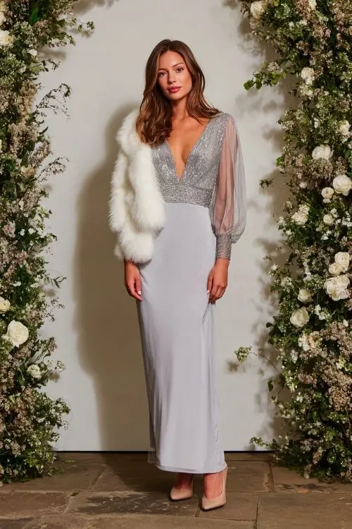 A woman over 30 wears a soft silver gown with full-length sheer sleeves adorned with beaded cuffs and a slight shimmer throughout the bodice