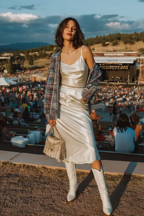 A woman over 30 wears sleek white cowboy boots with a satin midi skirt and fitted tank, layered with a plaid shirt worn open, accessorized with a fringe bag