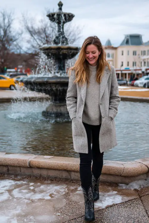A woman over 30 wears thermal black leggings with embroidered black cowboy boots, a soft gray turtleneck sweater, and a long wool coat
