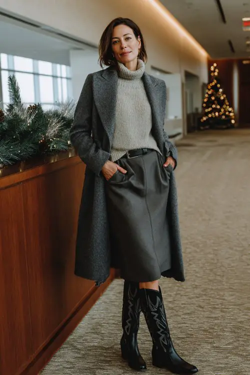 A woman over 40 wears a gray wool midi skirt, tall black cowboy boots, a tucked-in cashmere sweater, and a tailored wrap coat