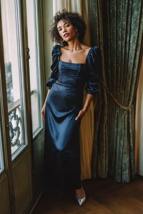 A woman over 40 wears a navy blue satin gown with ruched long sleeves, a square neckline, and silver pointed heels
