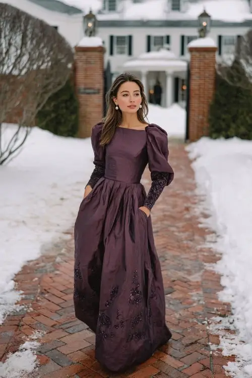 A woman over 40 wears a plum-colored column gown with tonal embroidery trailing down the sleeves and hemline, accessorized with pearl earrings