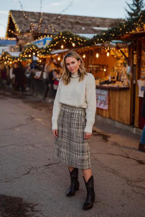 A woman over 40 wears a soft cream sweater tucked into a plaid midi skirt, paired with classic black cowboy boots