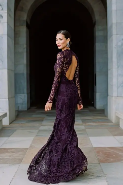 A woman walking through a grand archway in a dark plum long-sleeve gown with intricate lace detailing and a dramatic train, styled with a low bun and statement earrings