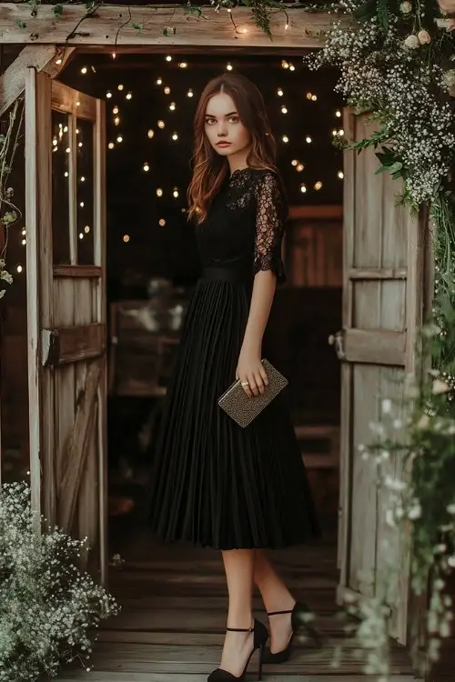 A woman wearing a black tea-length dress with lace sleeves and a soft pleated skirt, accessorized with heels and a clutch