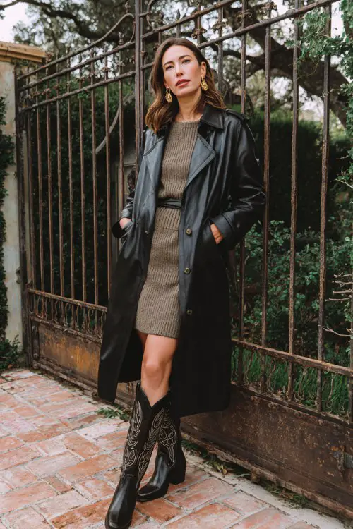 A woman wears a black faux-leather trench coat over a midi sweater dress in olive, paired with tall embroidered cowboy boots and statement earrings