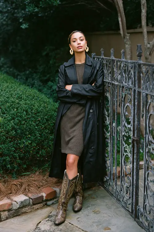 A woman wears a black faux-leather trench coat over a midi sweater dress in olive, paired with tall embroidered cowboy boots and statement earrings
