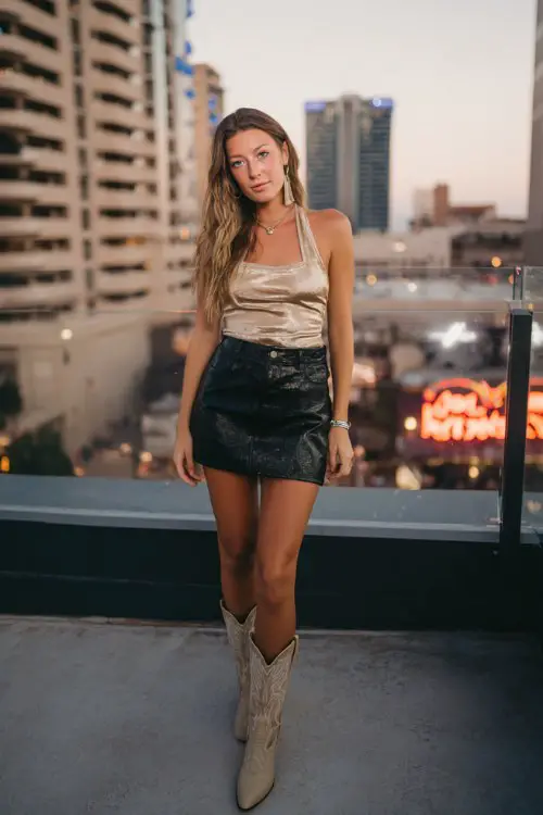 A woman wears a black leather mini skirt, a champagne satin halter top, and beige cowboy boots (3)