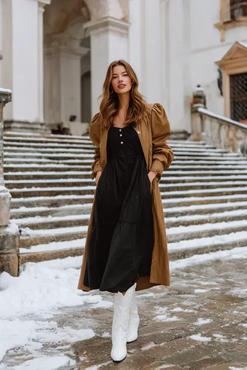 A woman wears a black midi dress with puff sleeves, a long camel trench coat, and tall white cowboy boots