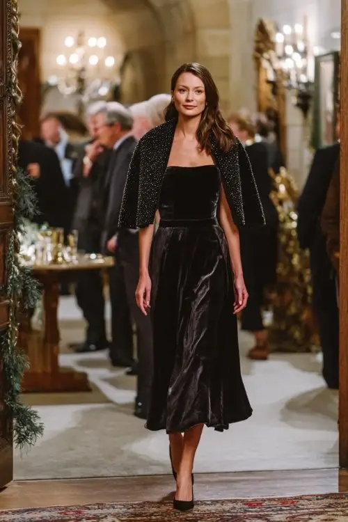 A woman wears a black velvet A-line dress with long mesh sleeves, paired with a pearl-embellished wool cape and sparkly pumps