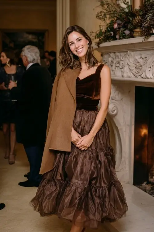 A woman wears a chocolate-brown organza gown with layered skirt and velvet waistband, layered with a camel wool cape coat and suede ankle-strap heels