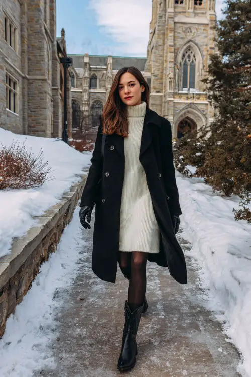 A woman wears a cream sweater dress with black tights and black cowboy boots, topped with a structured black overcoat and gloves