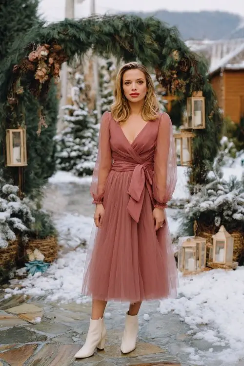 A woman wears a dusty rose tulle midi dress with long sleeves and a satin waist sash, paired with ivory ankle boots
