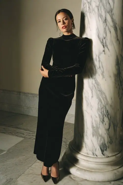 A woman wears a full-length black velvet gown with long fitted sleeves, a high neckline, and subtle crystal embellishments at the cuffs, styled with chandelier earrings