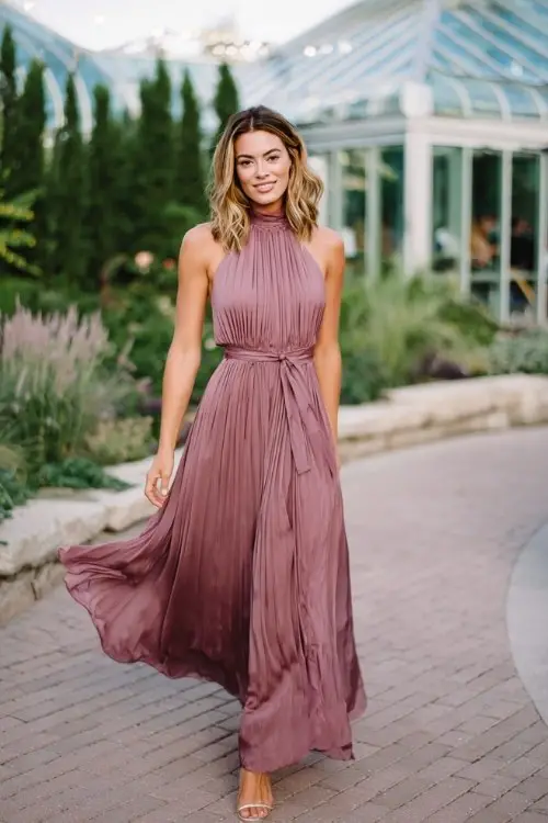 A woman wears a mauve satin high-neck gown with a pleated skirt and waist tie, layered under a flowing chiffon wrap that moves gently in the breeze