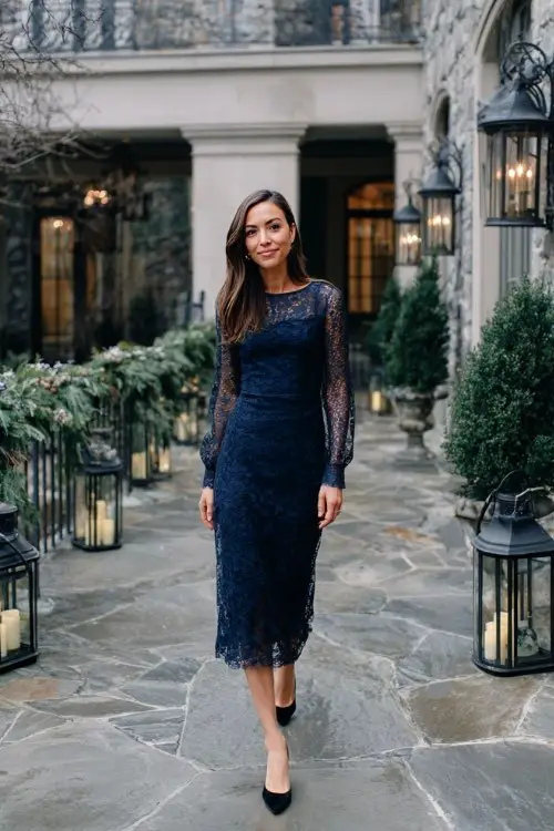 A woman wears a navy lace midi dress with sheer long sleeves and classic black pumps