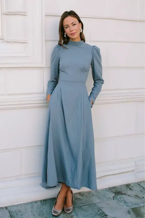 A woman wears a slate-blue gown with a high neckline, long tailored sleeves, and a gently flared hem, styled with pearl accessories and heeled pumps