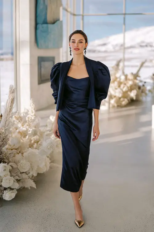 A woman wears a sleek navy satin slip gown under a structured taffeta puff-sleeve overlay jacket in matching tone, paired with statement earrings and gold kitten heels