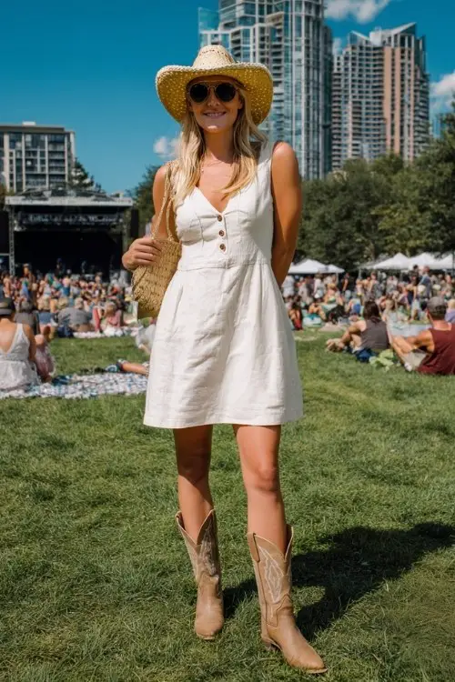 A woman wears a sleeveless white linen dress with beige cowboy boots and a straw cowboy hat for a sunny summer concert in the park