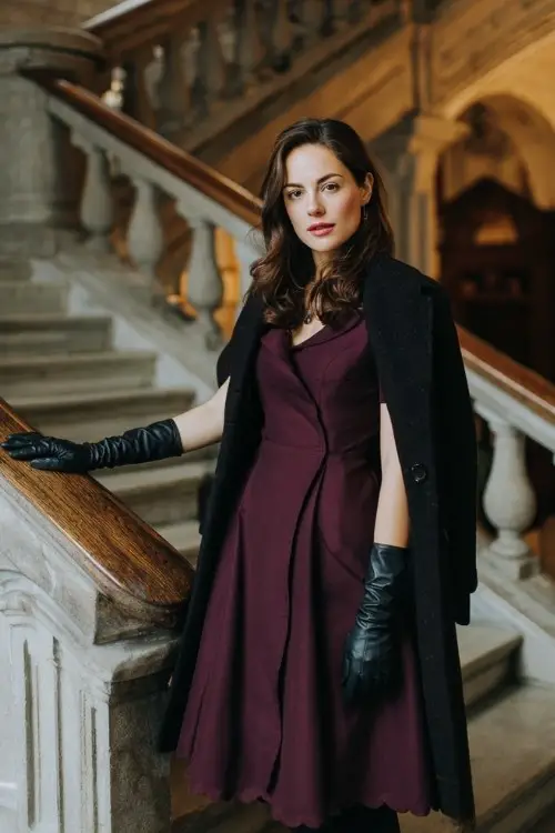 A woman wears a structured maroon midi dress with a scalloped neckline, styled with a charcoal wool swing coat, black tights, and leather gloves