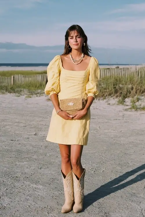A woman wears a yellow sundress with puff sleeves and beige cowboy boots, accessorized with a shell necklace and raffia clutch for a coastal summer wedding