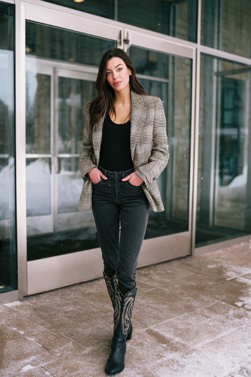 A woman wears black jeans with tall black cowboy boots, a mock-neck top, and a plaid blazer layered with a scarf