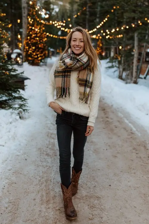 A woman wears dark skinny jeans tucked into short brown cowboy boots, paired with a cream cable-knit turtleneck and a plaid wool scar