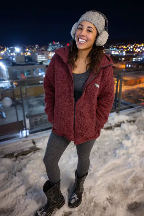 A woman wears gray leggings tucked into black cowboy boots, a warm maroon fleece jacket, and earmuffs