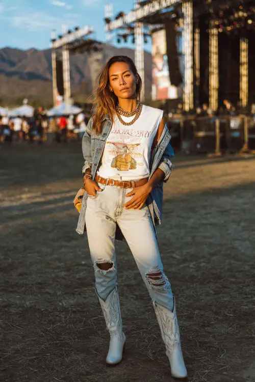 A woman wears white cowboy boots with high-waisted ripped skinny jeans, a fitted graphic band tee and oversized denim jacket, accessorized with layered chains and a mini crossbody bag