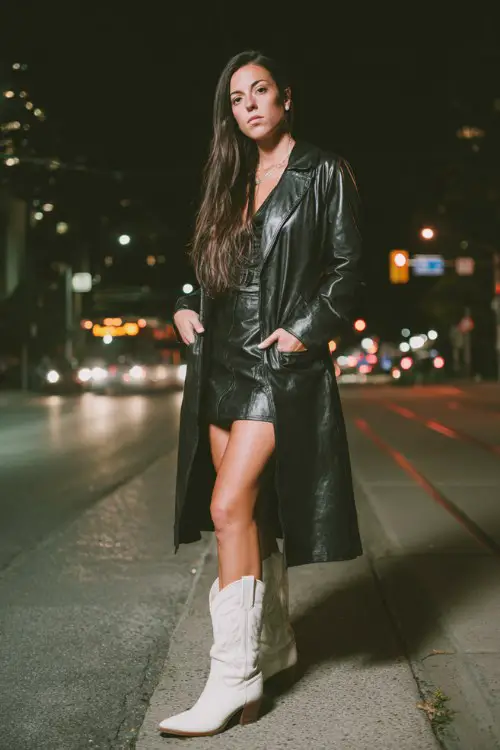 Bold fashion-forward woman at a nighttime urban concert wearing a long black leather coat over a fitted mini dress, white cowboy boots for a concert