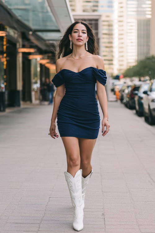 Elegant woman walking downtown before a concert, fitted navy off-shoulder mini dress, sleek silhouette, white cowboy boots for a concert, city street background, evening light