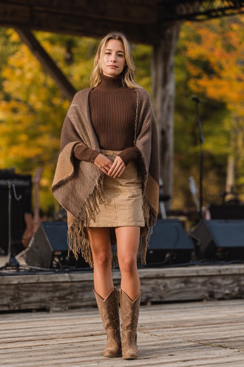woman in brown ribbed turtleneck sweater tucked into tan mini skirt, oversized colorblock poncho with fringe, tall brown leather cowboy boots
