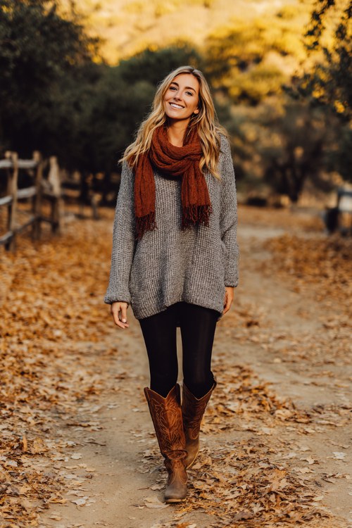 woman in oversized gray knit sweater, black leggings, rust-colored scarf, brown cowboy boots, walking on leaf-covered path