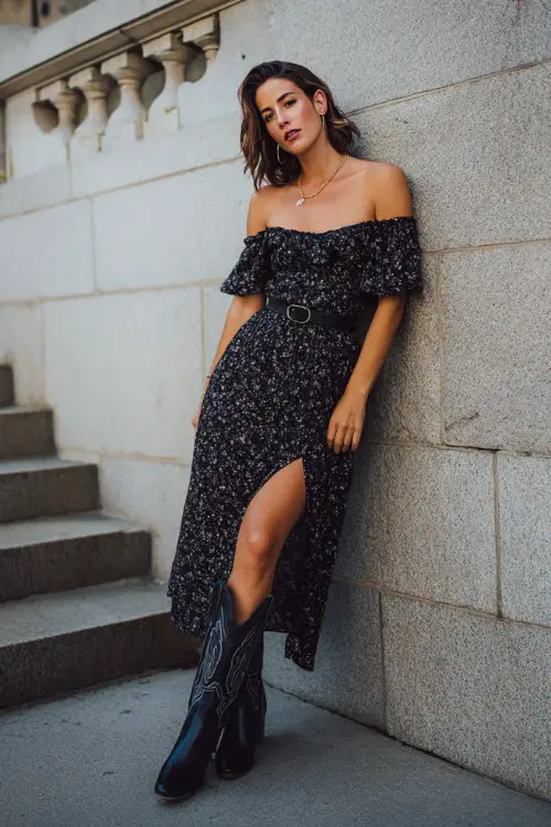 woman wearing black floral off-shoulder midi dress with slit, black leather cowboy boots, leaning against stone wall near stairs