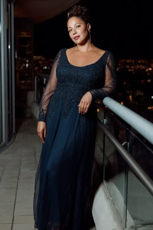A curvy woman wears a midnight blue beaded evening wedding gown with sheer sleeves, layered tulle hem and elegant floor-length silhouette