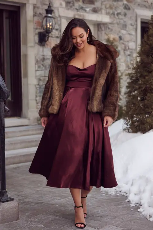 A curvy woman wears a satin midi dress in deep burgundy with a pleated skirt and square neckline, layered with a cropped faux fur jacket and ankle-strap heels