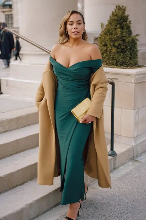 A curvy woman wears an emerald green off-the-shoulder gown with a camel wrap coat and gold clutch