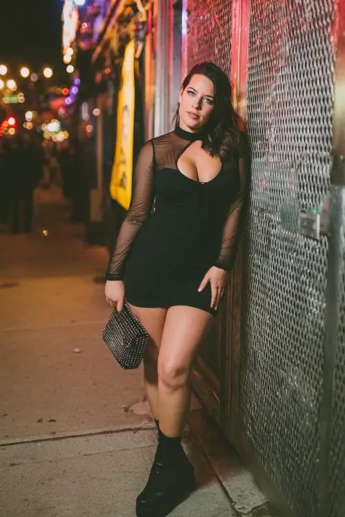 A plus size woman wears a black bodycon dress with sheer sleeves, platform boots, and a sparkling clutch