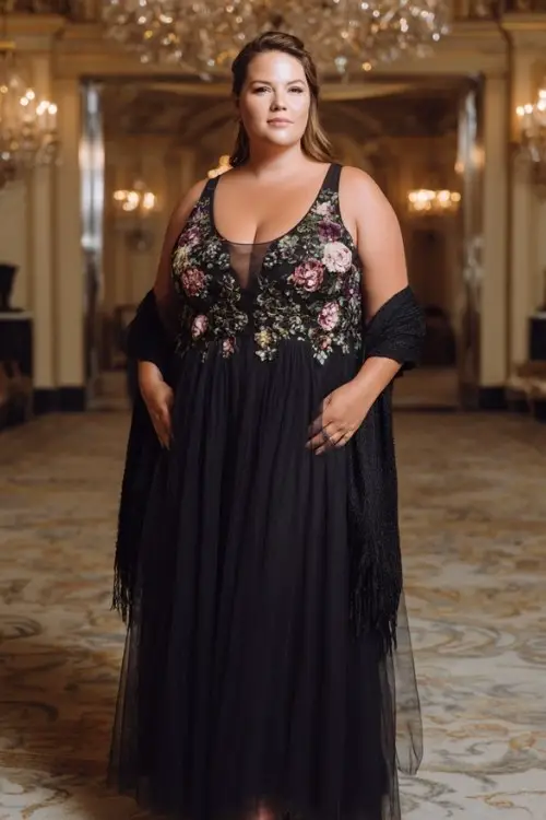 A plus-size woman wears a black tulle formal gown with floral appliqué details and illusion neckline, styled with metallic heels and elegant shawl