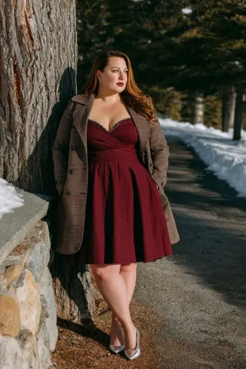 A plus-size woman wears a dark red A-line dress with chiffon sleeves and a beaded neckline, layered with a tailored wool coat and silver heels
