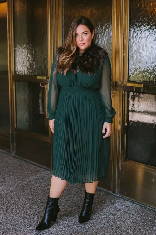 A plus-size woman wears a forest green pleated dress with sheer long sleeves, paired with black block-heel boots and a faux fur collar