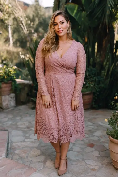 A plus-size woman wears a long-sleeve lace midi dress in dusty rose with elegant scalloped hem, paired with suede heels and delicate jewelry