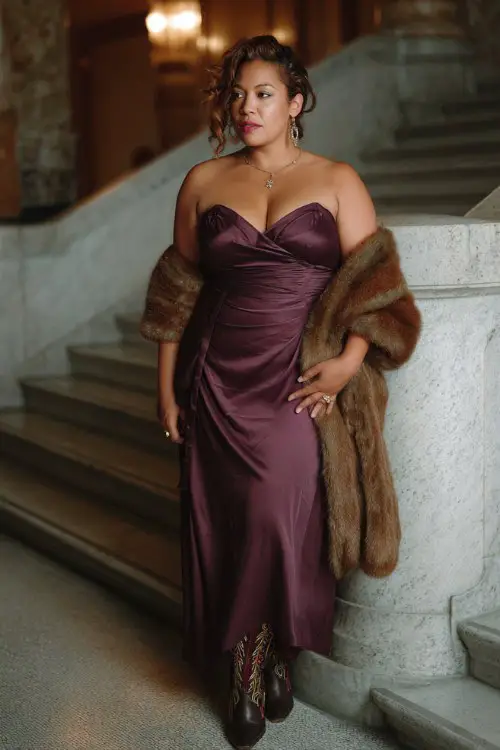 A plus size woman wears a plum satin evening dress with embroidered cowboy boots and a faux fur shawl