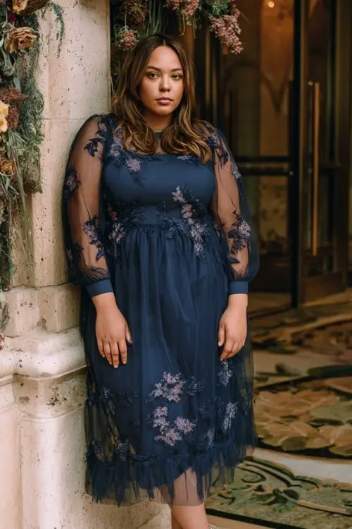 A plus-size woman wears a satin A-line formal dress in midnight blue with structured bodice and subtle shimmer overlay, styled with classic heels and tailored wool coat