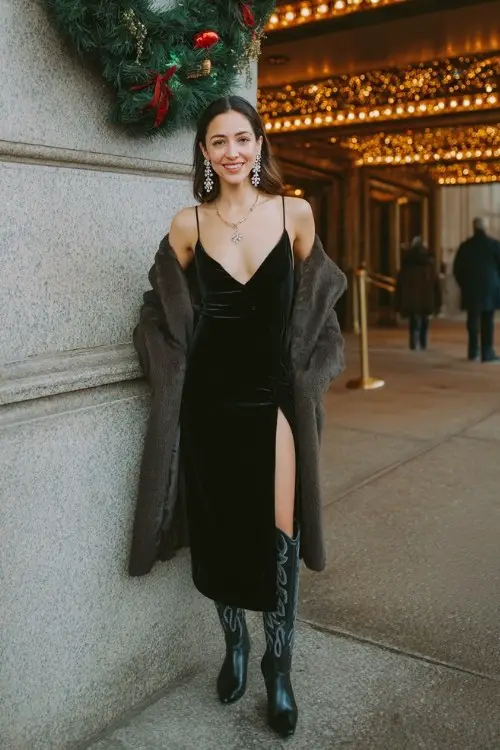 A woman in a black velvet midi dress with a deep neckline and crystal earrings, paired with tall black cowboy boots and a tailored wool coat