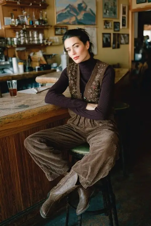 A woman in a cozy mountain café wears brown cowboy boots under corduroy wide-leg trousers, a turtleneck